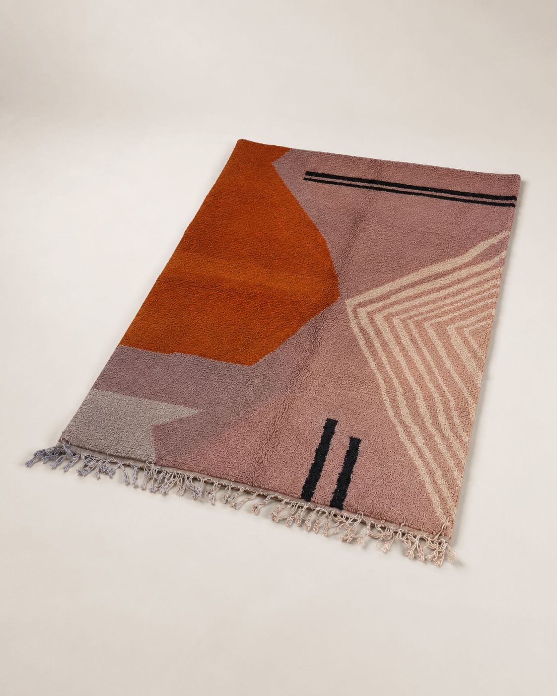A rectangular woven rug with a modern geometric pattern in dusty pink, rust orange, light gray, and cream, featuring fringed edges, photographed on a light beige studio background.